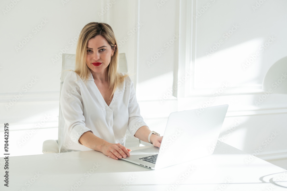 Delighted young blonde woman sitting at white table in blouse using laptop Delighted young blonde woman sitting at white table in blouse using laptop