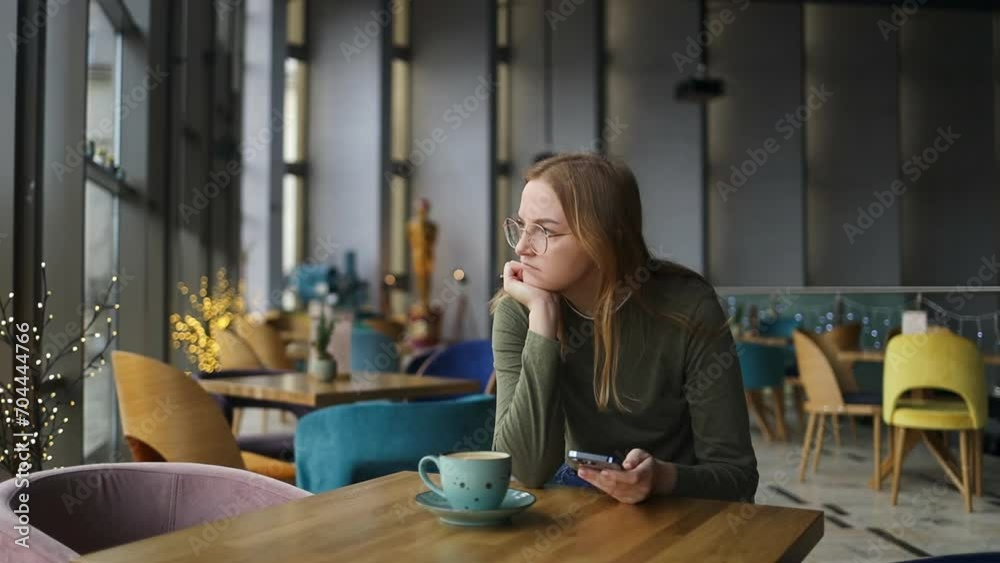 Woman sadly looking at smartphone in a cafe. Portrait of attractive beautiful sad lonely blonde female sitting near the window is missing, holds a smartphone in her hand. Girl getting surprising bad Woman sadly looking at smartphone in a cafe. Portrait of attractive beautiful sad lonely blonde female sitting near the window is missing, holds a smartphone in her hand. Girl getting surprising bad