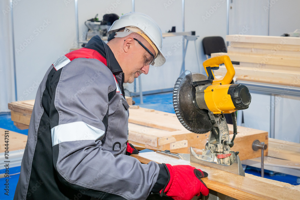 Man carpenter. Furniture production employee. Stationary circular saw near guy. Carpenter works in furniture shop. Woodworking worker. Man is carpenter in gray uniform. Guy working with circular saw Man carpenter. Furniture production employee. Stationary circular saw near guy. Carpenter works in furniture shop. Woodworking worker. Man is carpenter in gray uniform. Guy working with circular saw