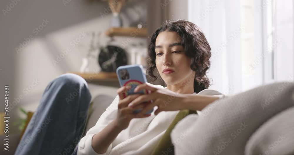 Girl uses smartphone while sitting on sofa in living room at home. Young girl sits on sofa, communicates with her loved one, smiles. A young smiling girl checks email, social networks, copy space.