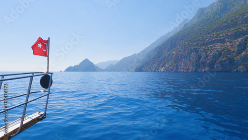 View of the bow of a Turkish flagged boat sailing in the blue sea along a beautiful rocky shore. Boat ride on the Mediterranean Sea. photo