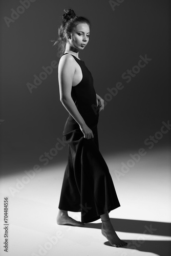 Black and white studio portrait of beautiful slender barefoot young woman in black dress full length