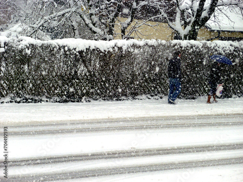 Frau mit blauem Schirm und der Mann hinter ihr gehen durch dicke Schneeflocken.  Wegen der dunklen Hecke sind sie kaum zu erkennen. Frische Fahrspuren im Schnee auf verschneiter Straße. Schneegestöber photo