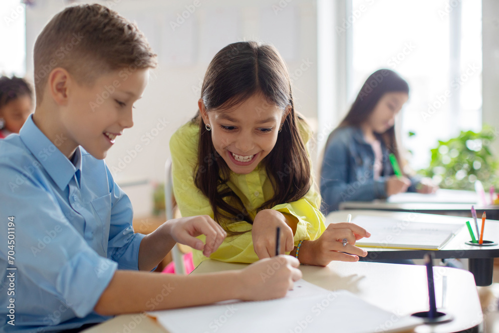 Schoolgirl sitting at table near boy classmate and pointing in copybook of friend with finger, enjoying learning process in classroom Schoolgirl sitting at table near boy classmate and pointing in copybook of friend with finger, enjoying learning process in classroom