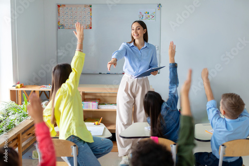 Friendly female teacher in classroom teaches elementary school students, kids raising hands to teacher aasks questions to small students sitting at their desks photo