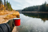 red mug with coffee or tea in hand on the background of the river in autumn