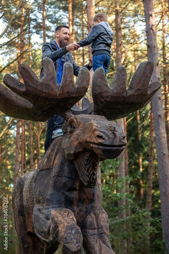 A father and son hold hands high on the antlers of a large wooden moose sculpture. Pine treetops in the background