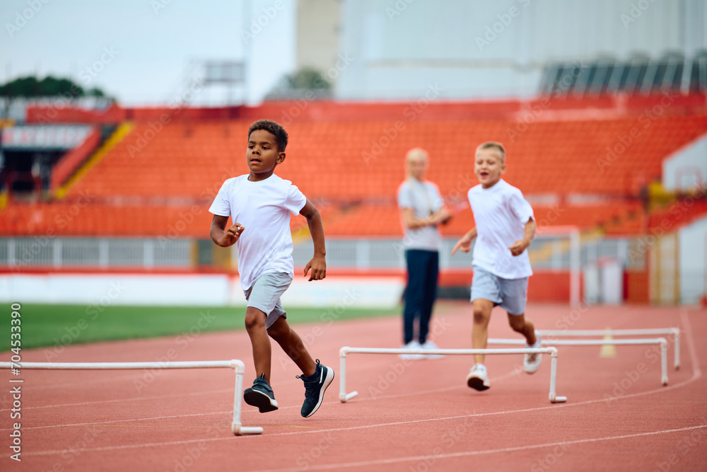 Black kid running on track with hurdles during sports training at stadium.