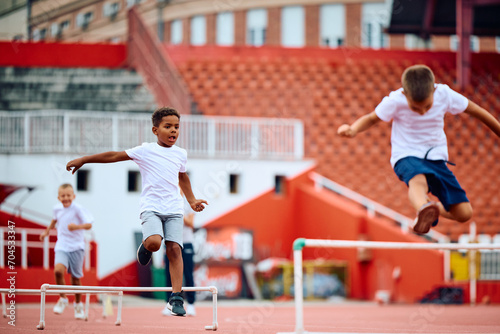 Little runners jumping over hurdles on running track at stadium.