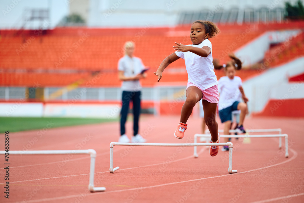 Black girl running on hurdling track at stadium.
