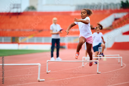 Black girl running on hurdling track at stadium.