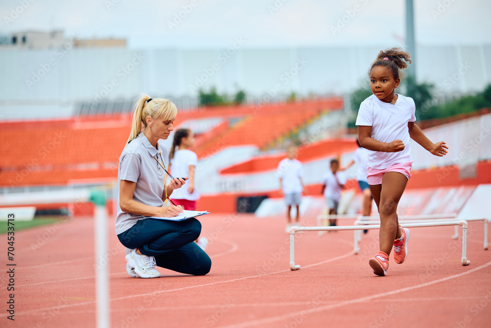 Sports teacher using stopwatch while black girl is jumping over hurdles on running track