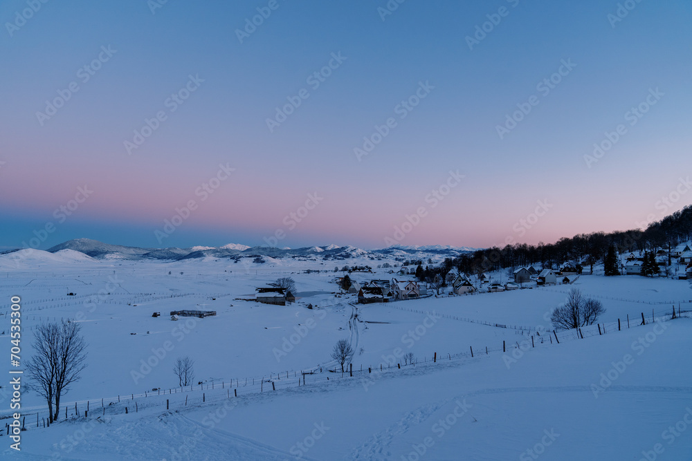 Pink-blue sunset over a snow-covered village on the edge of a forest in a mountain valley