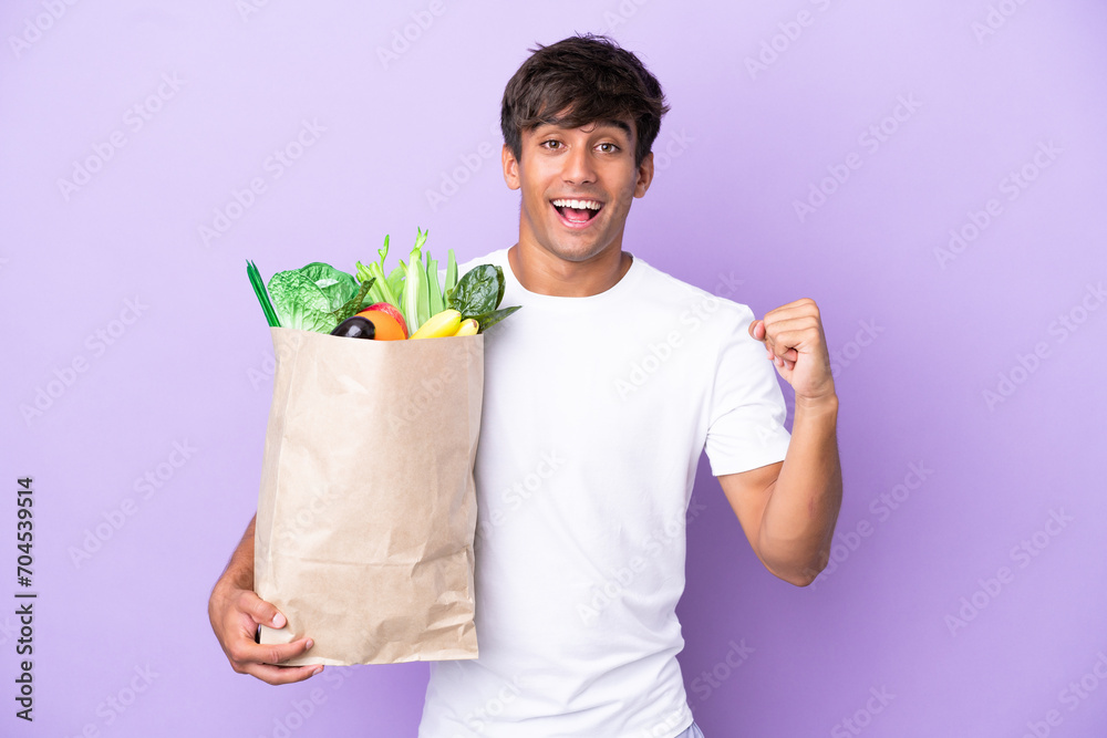 Young man holding a grocery shopping bag isolated on purple background celebrating a victory in winner position Young man holding a grocery shopping bag isolated on purple background celebrating a victory in winner position