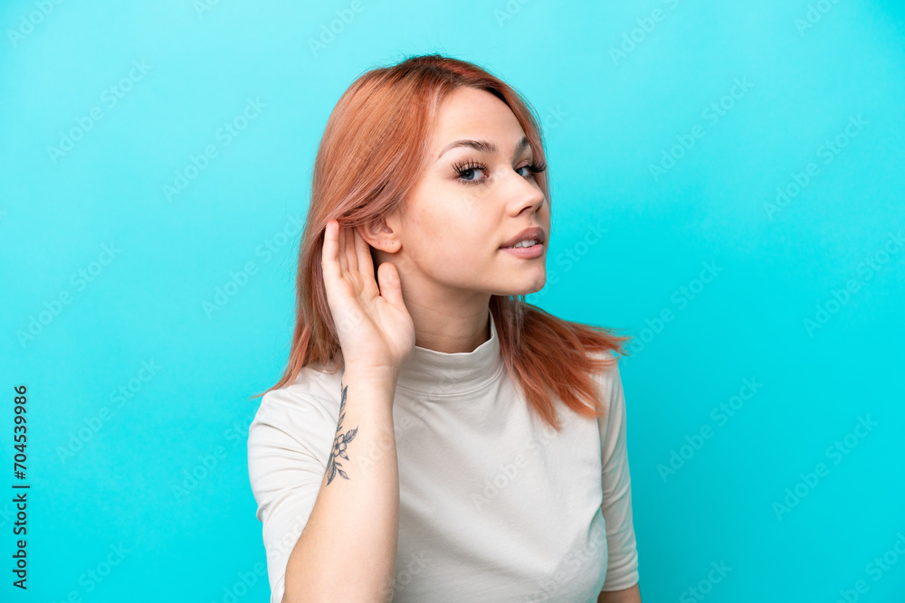 Young Russian woman isolated on blue background listening to something by putting hand on the ear