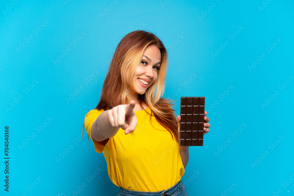 Teenager girl holding chocolat over isolated blue background pointing front with happy expression Teenager girl holding chocolat over isolated blue background pointing front with happy expression