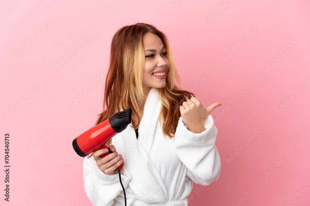 Teenager blonde girl holding a hairdryer over isolated pink background pointing to the side to present a product Teenager blonde girl holding a hairdryer over isolated pink background pointing to the side to present a product
