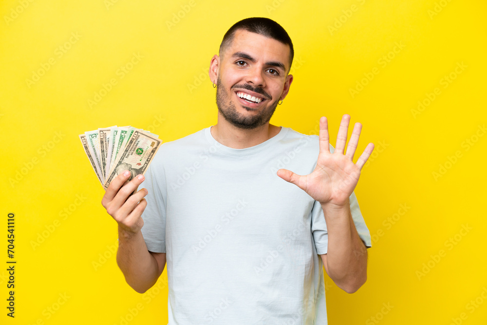 Young caucasian man taking a lot of money isolated on yellow background counting five with fingers Young caucasian man taking a lot of money isolated on yellow background counting five with fingers