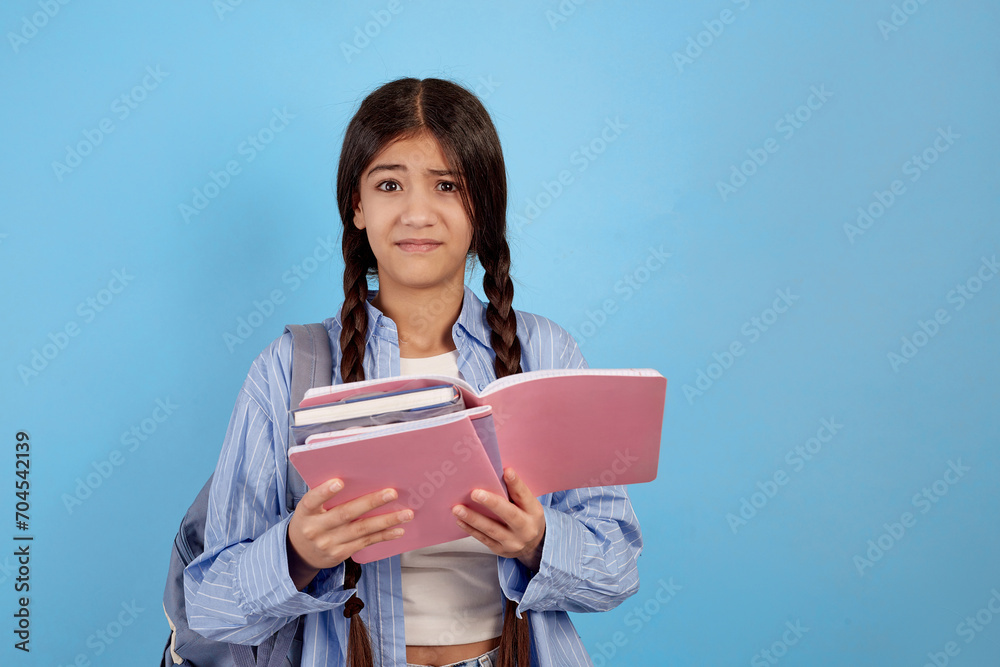Teenage girl books and notebooks with bag and looks dissatisfied on blue background. Teenage girl books and notebooks with bag and looks dissatisfied on blue background.