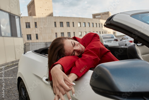 Happy young brunette sitting in convertible parked outdoors, leaning against car door