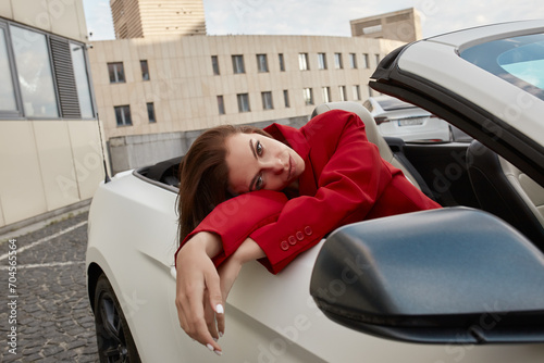 Young brunette in red blazer sitting in white convertible, leaning against car door