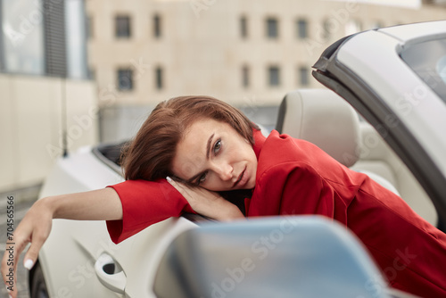 Attractive young brunette woman in red blazer sitting with head resting on hand in passenger seat of white convertible looking dreamily at camera
