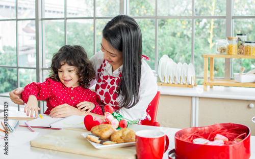 Beautiful Asian woman wearing apron and little cute daughter girl, giving gift and flowers on birthday or valentine day, smiling with happiness, having breakfast together,playing in kitchen at home.