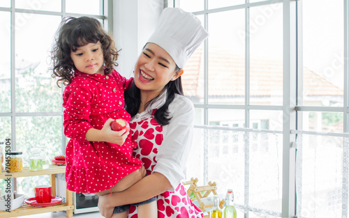 Beautiful Asian mother cooking for breakfast, carrying, taking care her mixed race adorable little daughter while sitting in cozy kitchen at home, happy smiling.