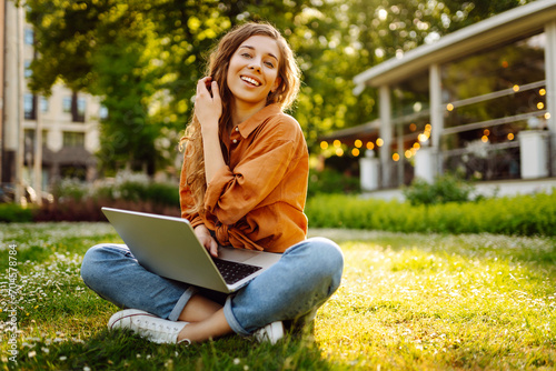 Cute woman spending time outdoors with laptop. Cheerful woman works online on the lawn in the park. Concept of freelancing, walking. photo