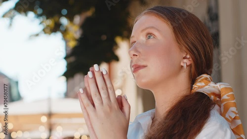 Portrait of red hair religion teenager girl praying closed eyes to God asking for blessing help forgiveness outdoors. Young woman clasping hands wishing luck in urban sunny city street. Town lifestyle photo
