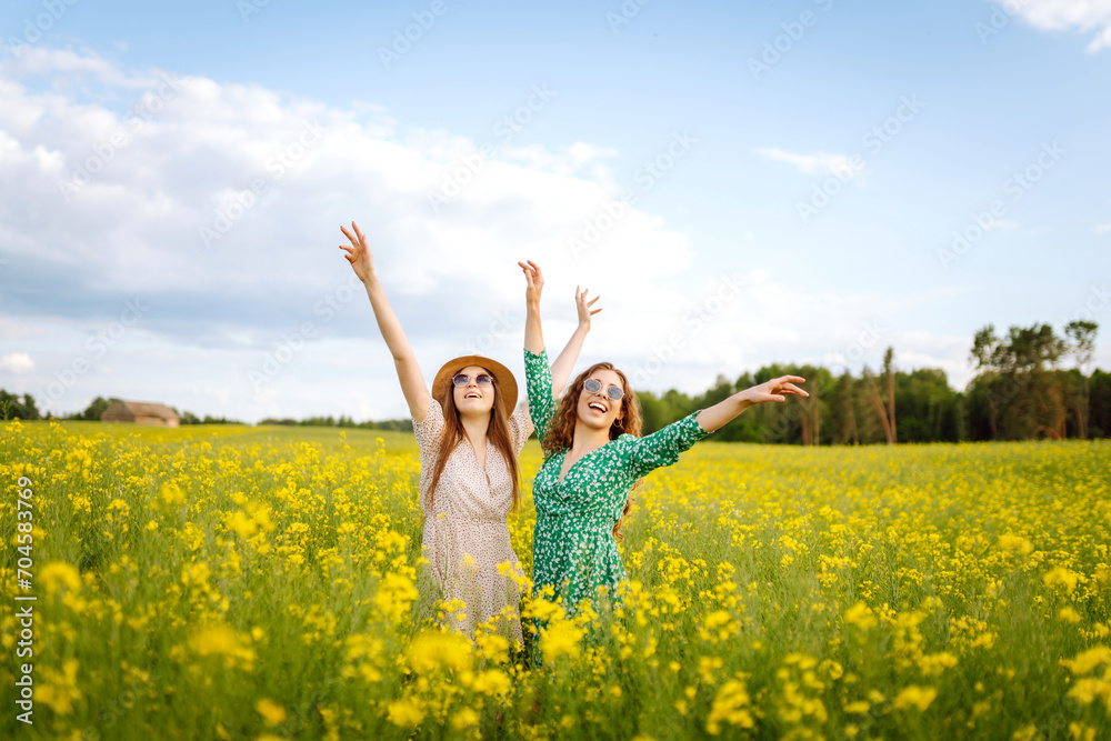 Two young women in beautiful dresses walk in a field with yellow blooming rapeseed. Beautiful girlfriends enjoying the weather, having fun in a flowering field. Concept of fun, relaxation. Two young women in beautiful dresses walk in a field with yellow blooming rapeseed. Beautiful girlfriends enjoying the weather, having fun in a flowering field. Concept of fun, relaxation.