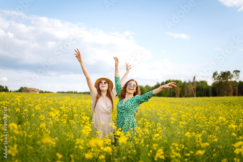 Two young women in beautiful dresses walk in a field with yellow blooming rapeseed. Beautiful girlfriends enjoying the weather, having fun in a flowering field. Concept of fun, relaxation.