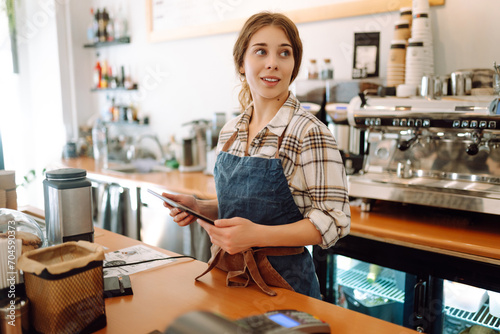 Portrait of a smiling cafe owner using a digital tablet behind the bar counter in a modern coffee shop. A young barista takes an order using a tablet. Small business concept. photo