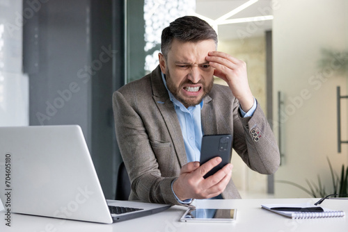 Angry and disappointed young male businessman sitting in the office at the desk and looking at the phone screen.