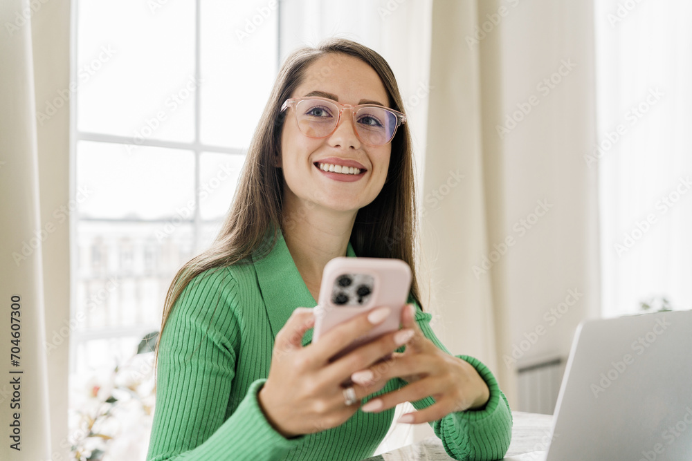 Smiling young woman in glasses using smartphone, cozy interior background with natural light. Smiling young woman in glasses using smartphone, cozy interior background with natural light.