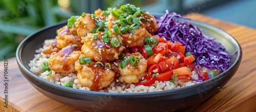 Close-up of a wooden table with a bang bang shrimp rice bowl, featuring fried shrimp, red cabbage, tomatoes, red pepper, and green onions, all topped with bang bang sauce. photo