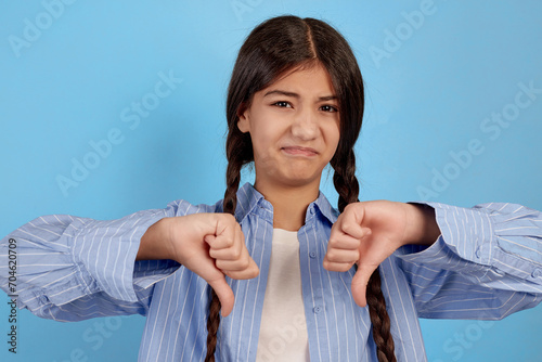 Malcontent teenage girl in blue shirt shows thumbs down on blue background. photo