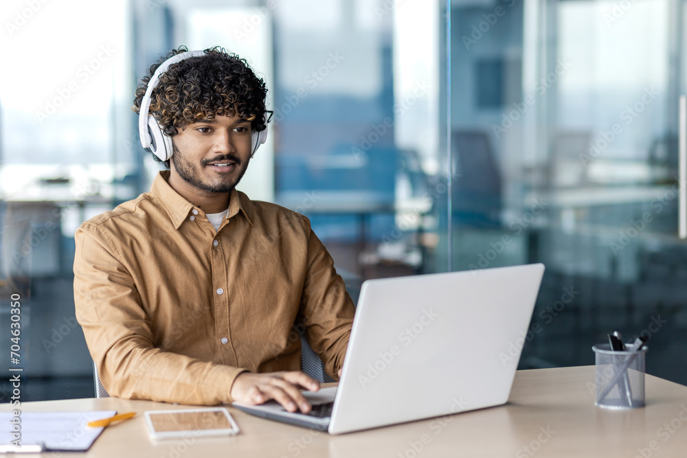 Young indian programmer working inside office with laptop, man in headphones coding new software, businessman developer satisfied with achieved results smiling. Young indian programmer working inside office with laptop, man in headphones coding new software, businessman developer satisfied with achieved results smiling.