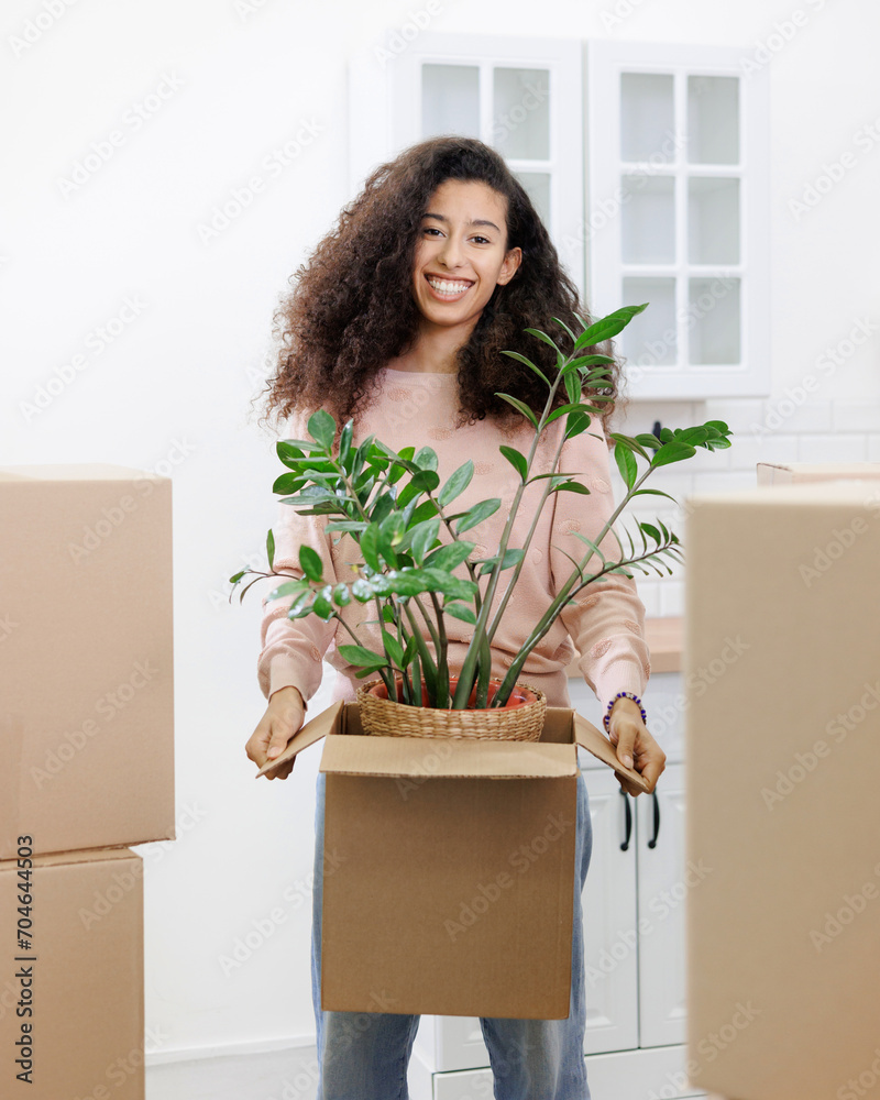Fototapeta premium happy young African American woman is moving into new home. person stands among cardboard boxes and smiles, holding a houseplant, mortgage or rental property in his hands. mover service for moving.