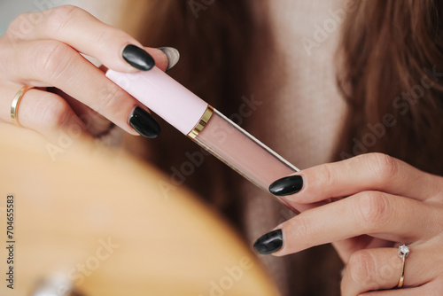Long haired woman opening lip gloss. Cosmetics background. Pink lips. Girl doing makeup in front of a mirror. Closeup on product container in tube. photo