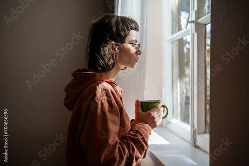 Lonely sad teenager with cup of tea looking a window, brooding. Unsociable teen girl spending weekend at home because of friendlesness, social difficulties, problems in communication, misunderstanding