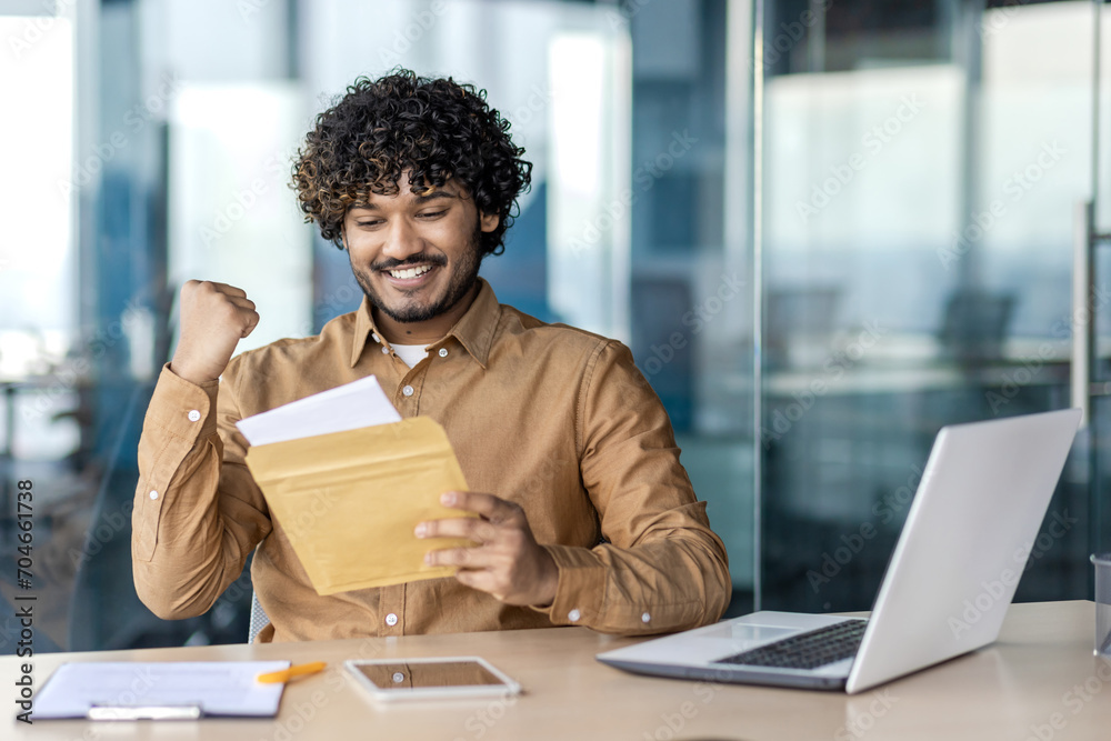 Businessman received postal envelope notification letter with good news, man celebrates holding hand up, joyfully reading news, sitting inside office with laptop. Businessman received postal envelope notification letter with good news, man celebrates holding hand up, joyfully reading news, sitting inside office with laptop.