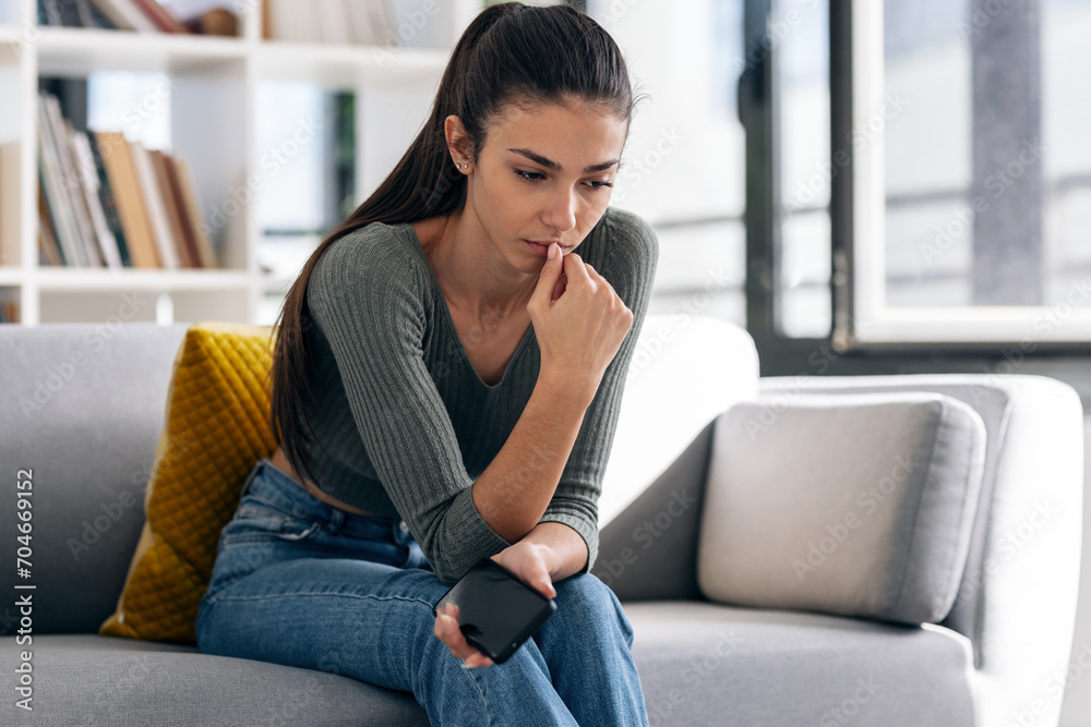 Depressed young woman thinking about her problems while holding mobile phone sitting on the sofa at home.