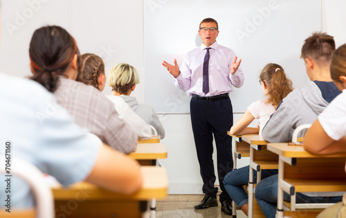 Teacher explaining studying material to teenagers during lesson at school, standing at whiteboard