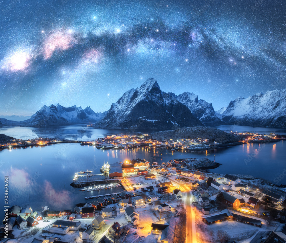 Milky Way over the snowy village, islands, rorbuer, city lights, sea, mountain, starry sky at winter night. Landscape with town, street illumination, rocks. Reine, Lofoten islands, Norway. Aerial view