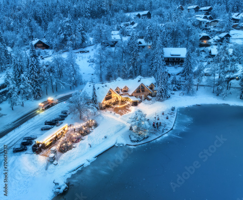 Aerial view of illuminated houses in fairy village in snow, forest, Jasna lake, street lights at winter night. Top view of alpine countryside, snowy pine trees, road at dusk. Kranjska Gora, Slovenia