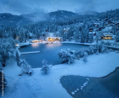 Aerial view of fairy alpine village in snow, forest, Jasna lake, houses, street lights at winter night. Top view of mountains in fog, illumination, snowy pine trees at dusk. Kranjska Gora, Slovenia
