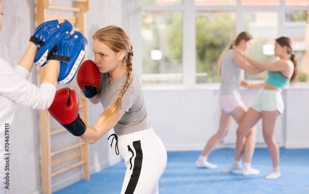 Obraz premium Concentrated sporty teen girl in boxing gloves practicing self-defense techniques in gym, throwing punches on focus mitts in hands of instructor..