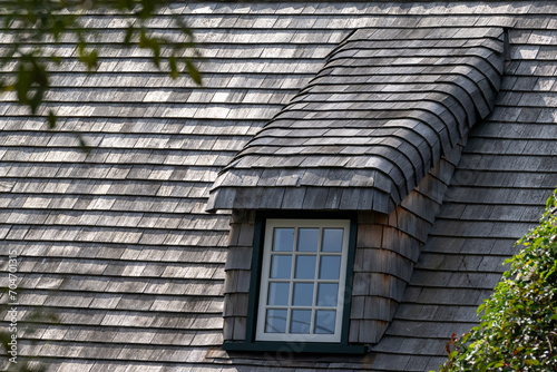 A steep roof of a vintage building with cedar wood shingles. There's a long dormer covered in weathered wood. The frame of the window is green in color. It has multiple glass panes with white trim. photo
