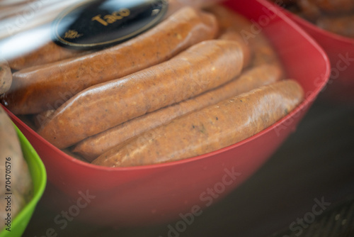A red plastic bowl filled with taco sausages in a display case with a black sign and gold letters. The uncooked Mexican protein is raw. The stuffed ground meat weiners are for sale at a market.  photo